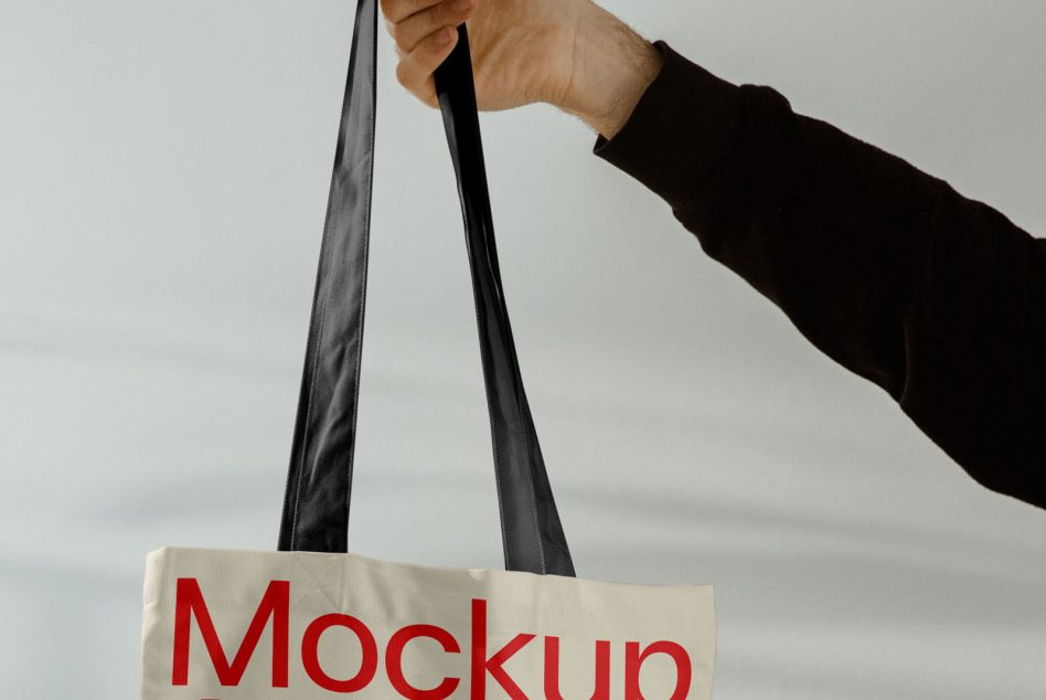 Hand holding cream canvas tote bag with black handles, red Mockup text visible