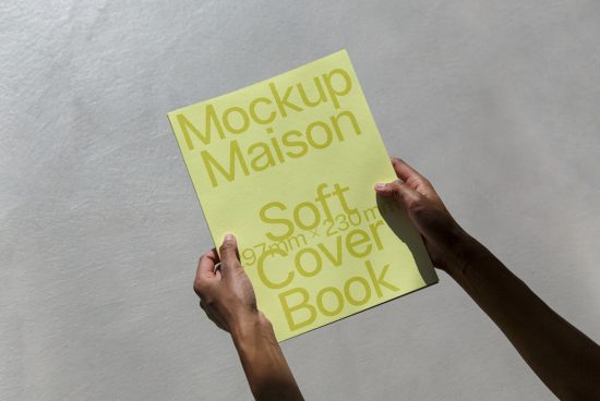 Person holding yellow softcover book mockup with sunlight shadow texture, ideal for book cover design presentation.