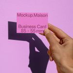 Hand holding a pink business card mockup with shadow on a textured purple wall, design presentation, 85 x 55 mm size model.