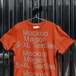 Orange t-shirt mockup on hanger with text graphics against metallic ribbed background, ideal for apparel design presentations.