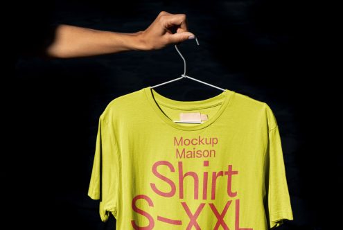 Hand holding hanger with lime green T-shirt mockup, graphic design display, dark backdrop, clothing template, apparel presentation.