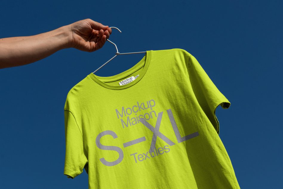 Person holding bright neon green t-shirt mockup on hanger against blue sky, clear for graphic design and branding display.