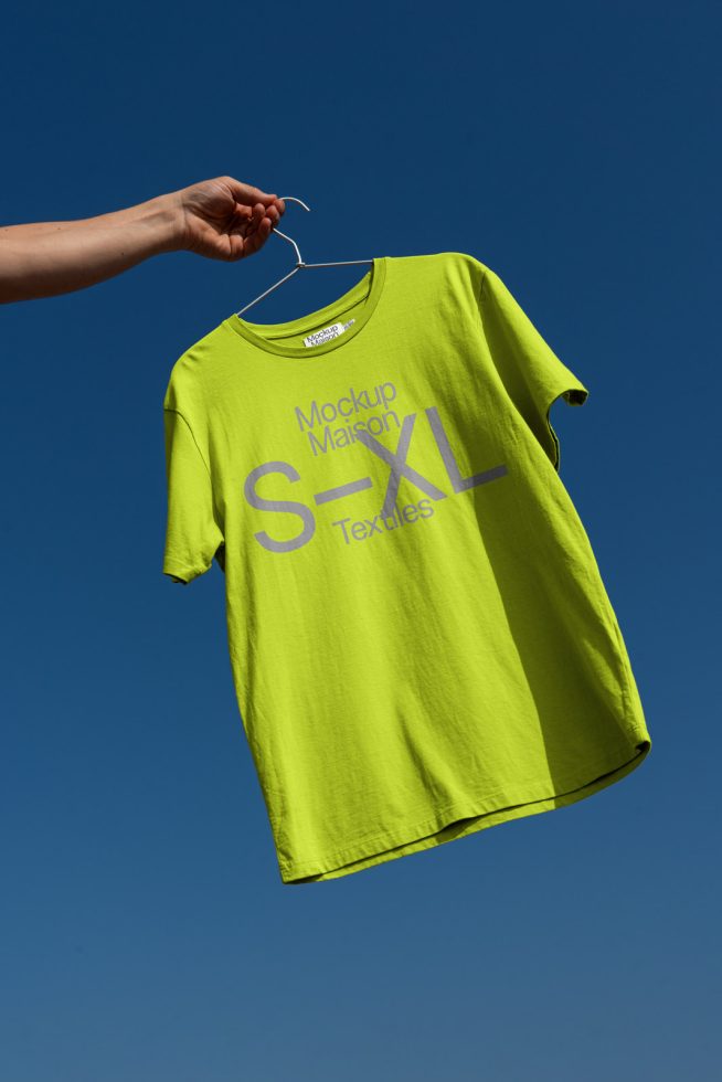 T-shirt mockup with a blue sky background, hand holding hanger, vibrant neon green textile, clear space for design.