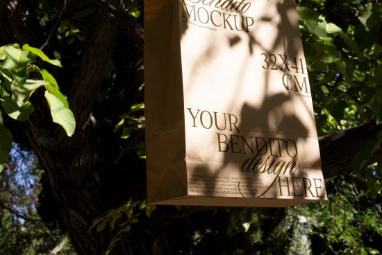 Paper bag mockup hanging on a tree in sunlight, displaying space for design placement, ideal for eco-friendly packaging design presentation.
