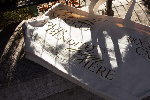 Elegant tote bag mockup with shadow overlay and script typography design, laid on outdoor ground in sunlight, ideal for branding presentation.