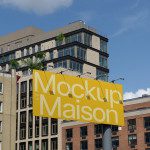 Urban billboard mockup displaying yellow sign with text 'Mockup Maison' against a backdrop of buildings, perfect for designers to showcase advertising designs.