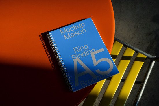 A5 notebook mockup with ring binding on orange surface and metal chair, showcasing design presentation, spiral notebook graphic.
