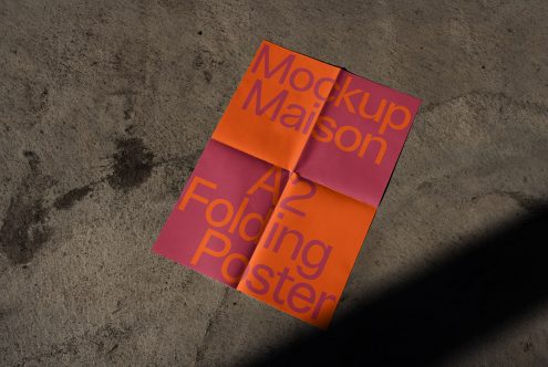 A2 size folded poster mockup in orange and pink with typography, resting on a textured concrete floor in sunlight for design presentation.