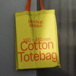 Green cotton tote bag mockup with red text hanging on a wall, realistic design asset for branding and packaging.