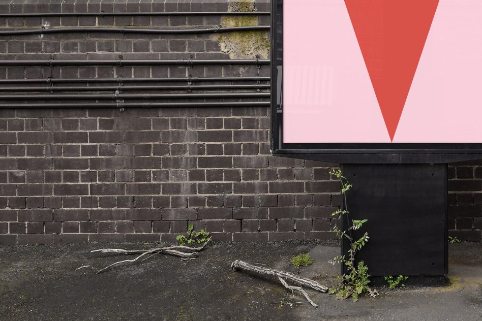 Red V-shape graphic on pink billboard against dark brick wall with weeds