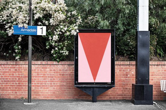 Outdoor advertising mockup of a billboard with red and pink design at a train station, surrounded by lush foliage and brick wall.
