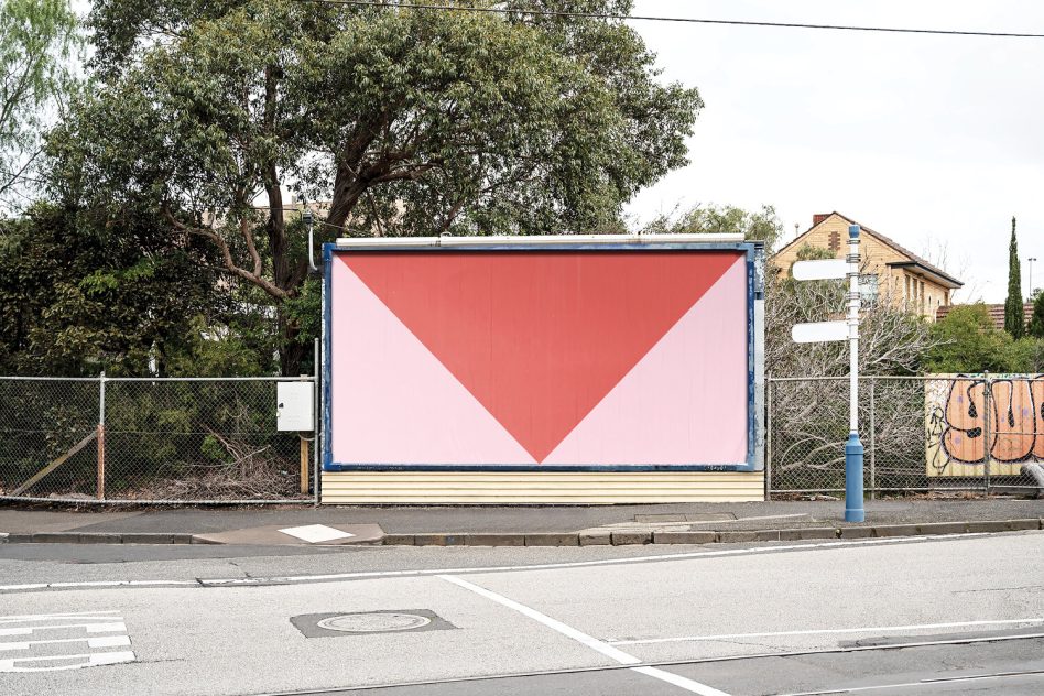 Street billboard with red downward triangle on light pink, brick house visible behind fence