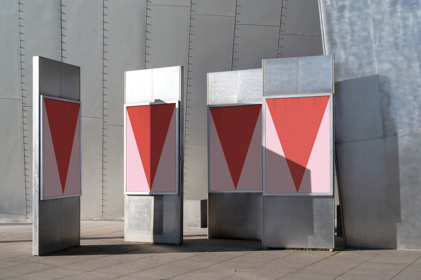 Outdoor advertising mockup of three vertical billboards with red and pink geometric shapes, against a modern gray building wall for design display.