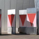 Outdoor advertising mockup of three vertical billboards with red and pink geometric shapes, against a modern gray building wall for design display.