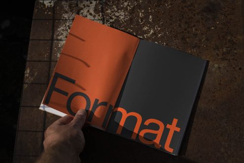 Open magazine mockup on a textured surface with hand holding one page, showcasing bold font typography design.