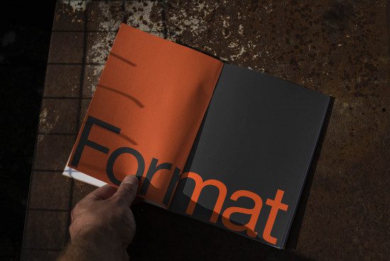 Open magazine mockup on a textured surface with hand holding one page, showcasing bold font typography design.