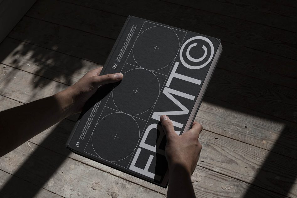 Person holding a black and white printed design portfolio with geometric graphics, showcasing modern typography mockup on wooden floor.