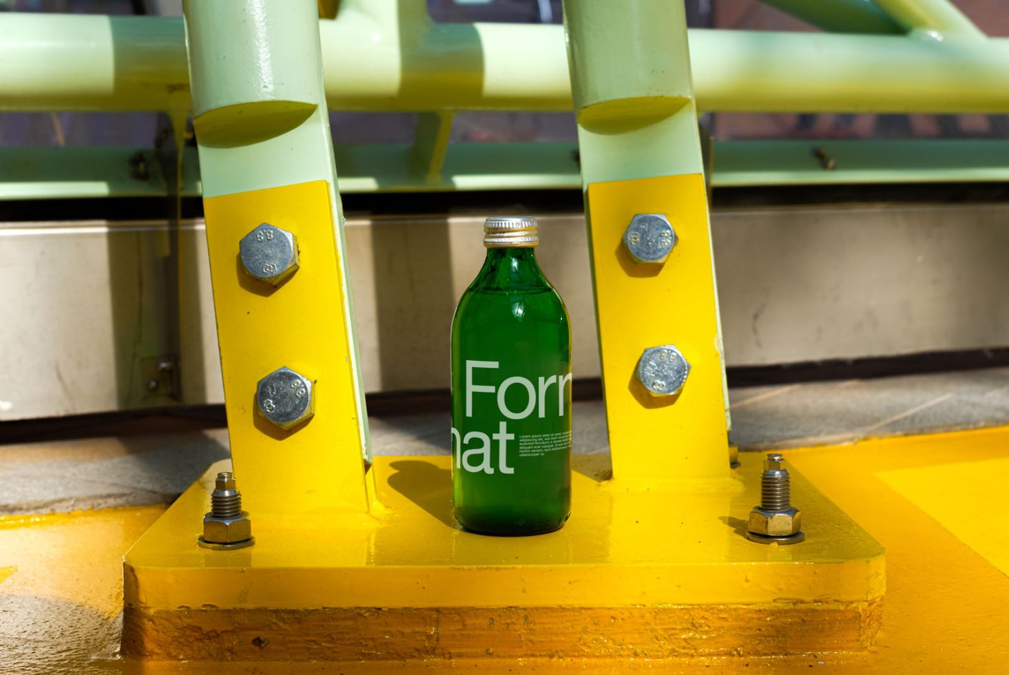 Green glass bottle with label design mockup on yellow base between industrial pipes, showcasing product packaging presentation for designers.