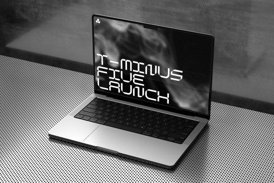 Laptop on a perforated metal table displaying typography design T-minus Five Launch, ideal for mockup graphics, product presentation.