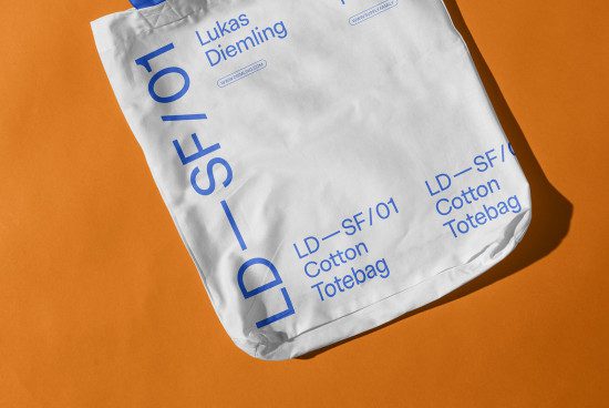 White cotton tote bag mockup with blue typography design, lying on an orange background for graphic design assets.