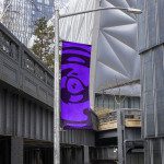 Urban street banner mockup hanging from a metal pole with a playful purple character design, showcasing outdoor advertising in a modern city setting.