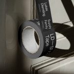 Roll of black duct tape with typography mockup casting shadow on white wall, useful for graphic design presentations.