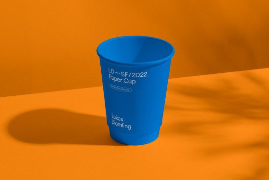 Blue paper cup mockup on orange background displaying text and website URL, ideal for branding and packaging design presentations.