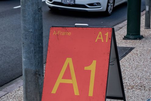 Red A-Frame sign with yellow A1 text on sidewalk, street sign, car in background, urban environment, realistic mockup for signage design.