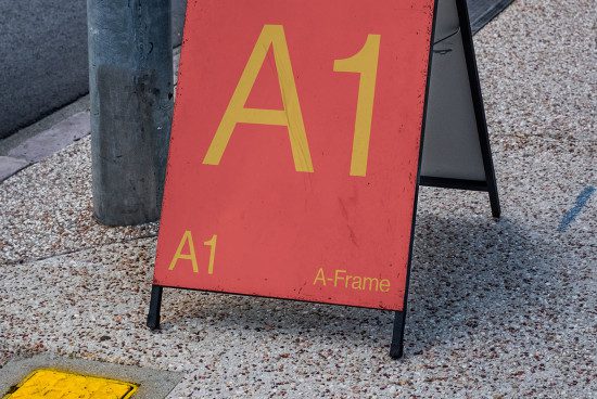 Red and yellow A-Frame sign mockup on a sidewalk, showcasing bold typography and weathered texture, perfect for brand display designs.