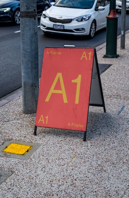 Red A-frame sign mockup on a city sidewalk with clear space for design presentations, background includes cars and street elements.