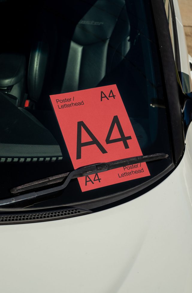 A4 red paper mockup for poster or letterhead design placed under windshield wiper on a car, creative outdoor presentation.