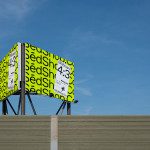 Billboard mockup with bright yellow background and bold black text, perched above a grey building against a clear blue sky, ideal for designers.