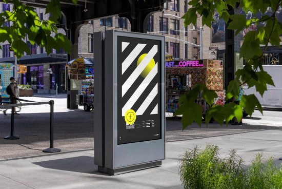 Urban outdoor advertising kiosk mockup with black and yellow geometric design in a dynamic city environment surrounded by greenery.
