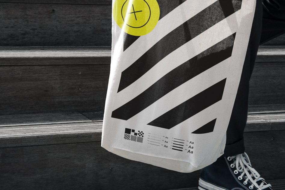 Tote bag mockup with bold black and white diagonal stripe design, typographic elements, and person holding it against a dark textured background.