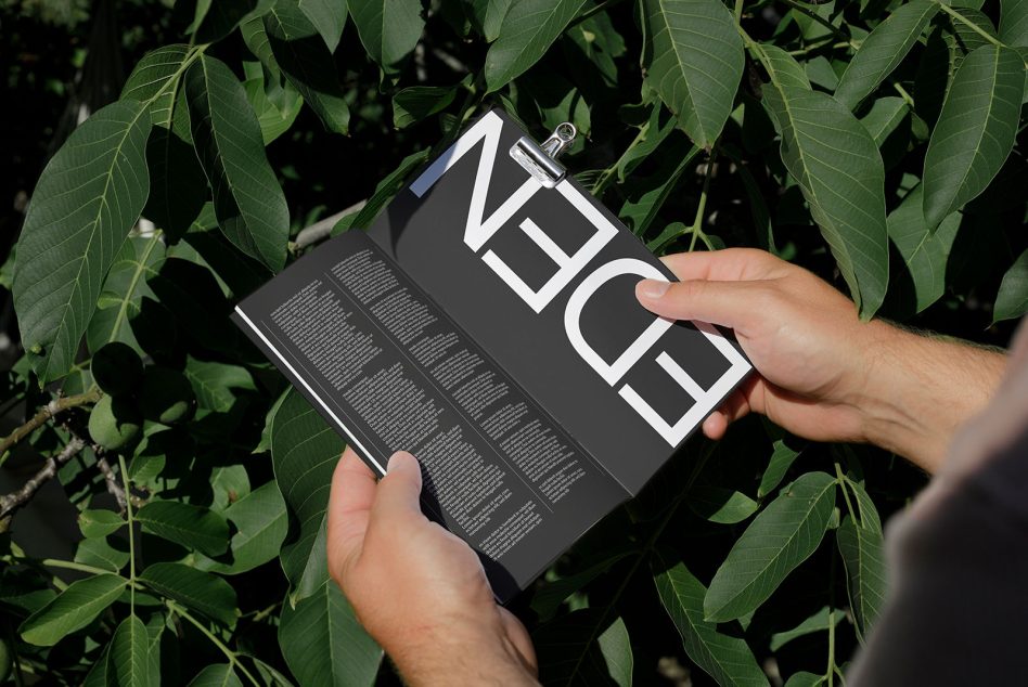 Person holding open magazine with bold design mockup among green leaves, showcasing clear typography and layout design.