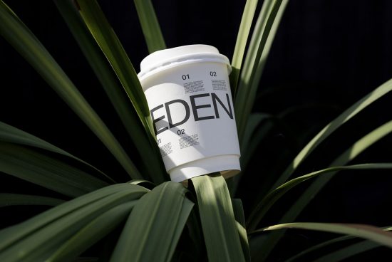 Disposable coffee cup with modern typography mockup resting on green plant leaves, ideal for eco-friendly branding presentation.