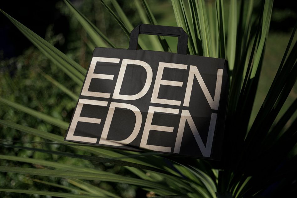 Black tote bag with cream EDEN text stacked, surrounded by green plant leaves