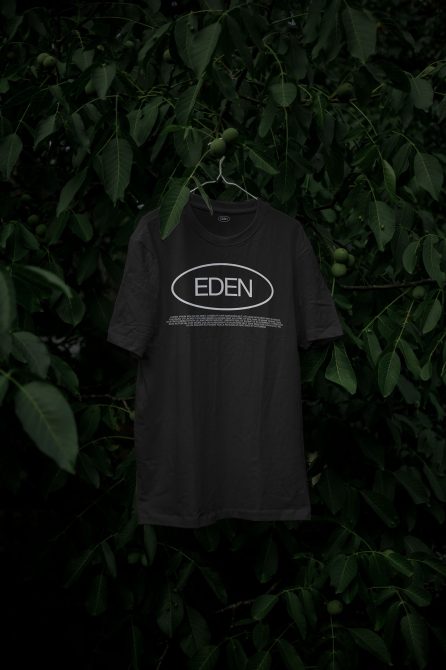 T-shirt mockup hanging on a hanger amidst green leaves, displaying a black shirt with 'EDEN' logo, ideal for clothing design presentations.