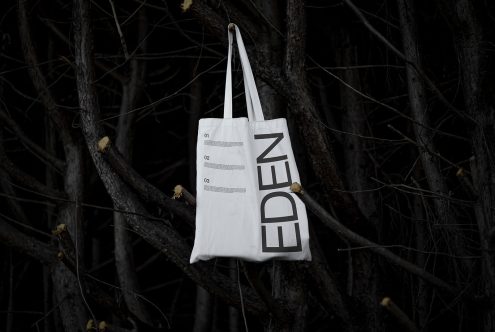 White tote bag with bold Eden text design mockup hanging on dark tree branches, stark contrast, ideal for showcasing typography and branding designs.