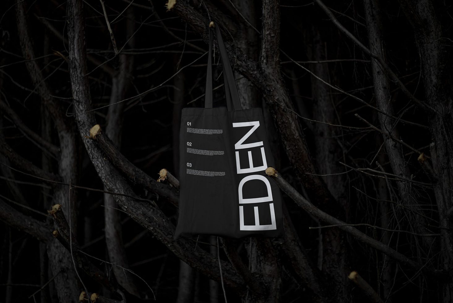 Dark tote bag mockup with EDEN text hanging on tree branches, showcasing design for digital asset marketplace, targeted at designers.