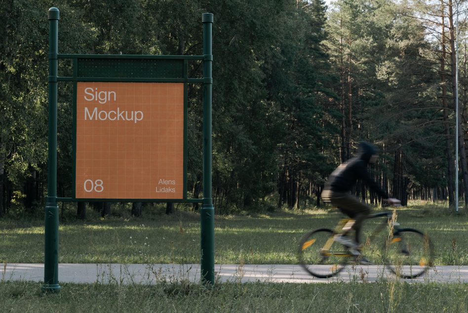 Outdoor sign mockup with orange panel on green metal frame, cyclist riding past