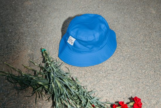 Blue bucket hat lying on concrete ground beside greenery and red flowers, fashion mockup for design presentation.