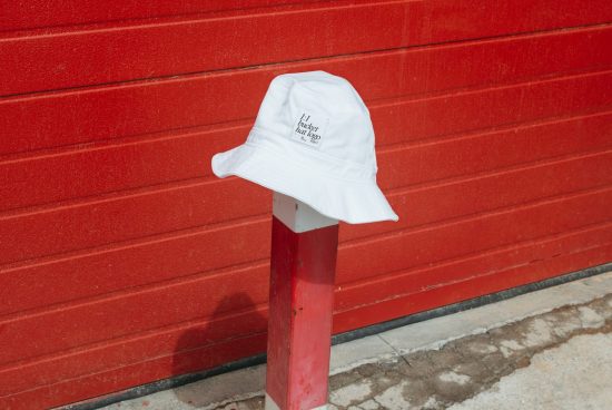 White bucket hat with logo mockup on post against red wall, urban streetwear design, fashion accessory graphic display.
