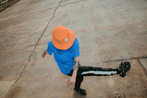 Person in dynamic urban fashion pose wearing bright orange bucket hat, blue t-shirt, and black pants suitable for streetwear design mockups.