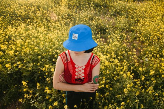Person in blue bucket hat and red top standing in a yellow flower field, fashion mockup with copyspace, apparel design showcase.