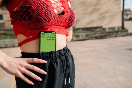 Woman with red fishnet top and black pants holding a smartphone with green screen mockup for design display.