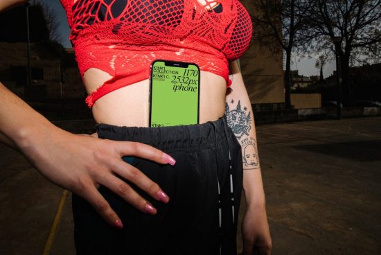 Woman with smartphone mockup, urban style clothing, tattoo visible, showcasing screen for design presentation.