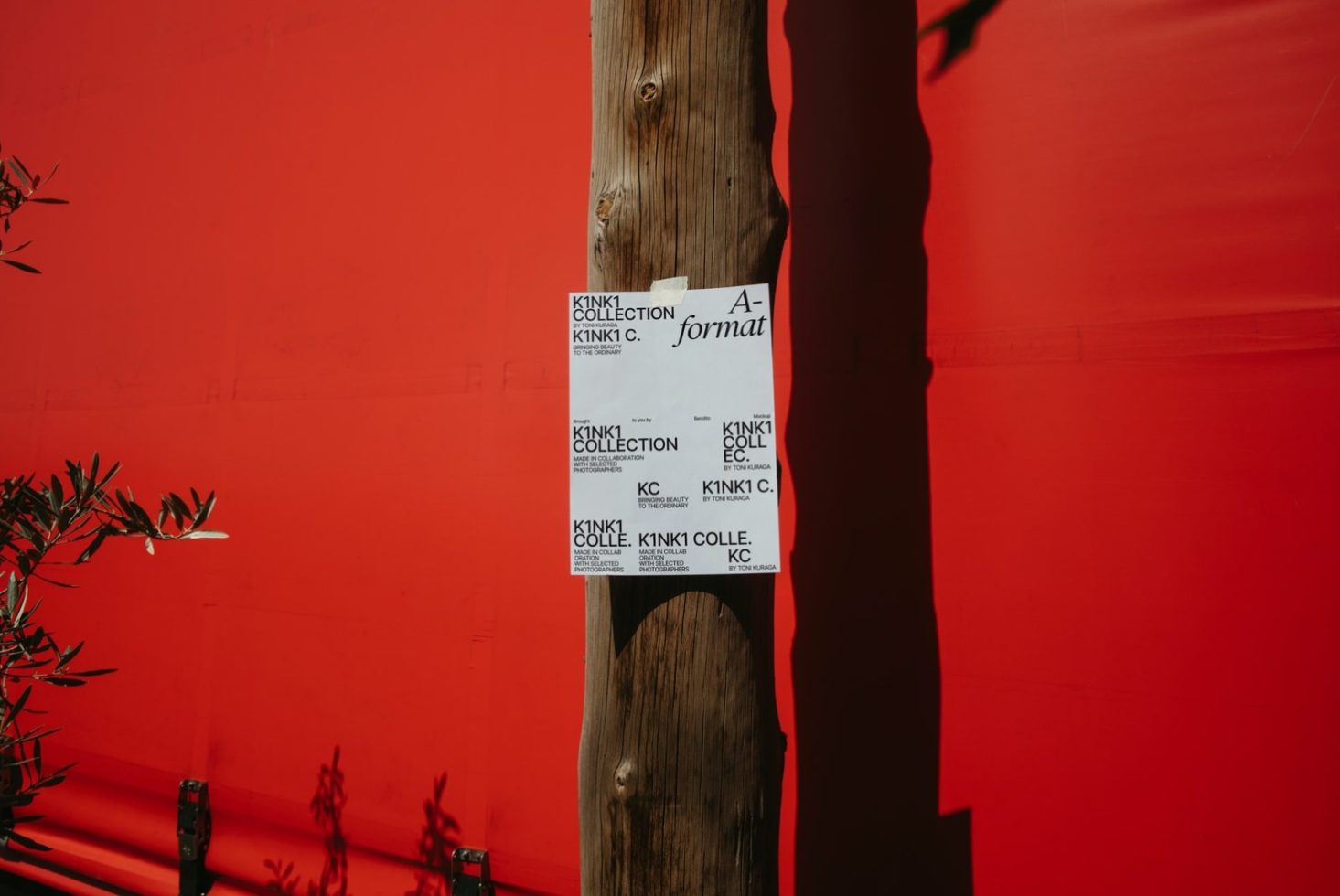 Bright red wall with stark shadow, wooden pole with attached paper mockups featuring black text, graphic design presentation, artsy urban scene.