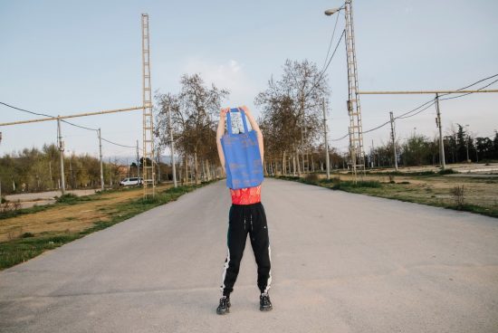 Person with bag over head stands on empty road, quirky streetwear mockup, outdoor urban setting, designer asset.
