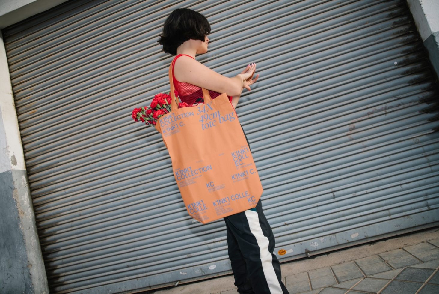 Woman with floral dress and tote bag mockup against metal shutter for graphic design assets, showcasing vibrant typography and street style.